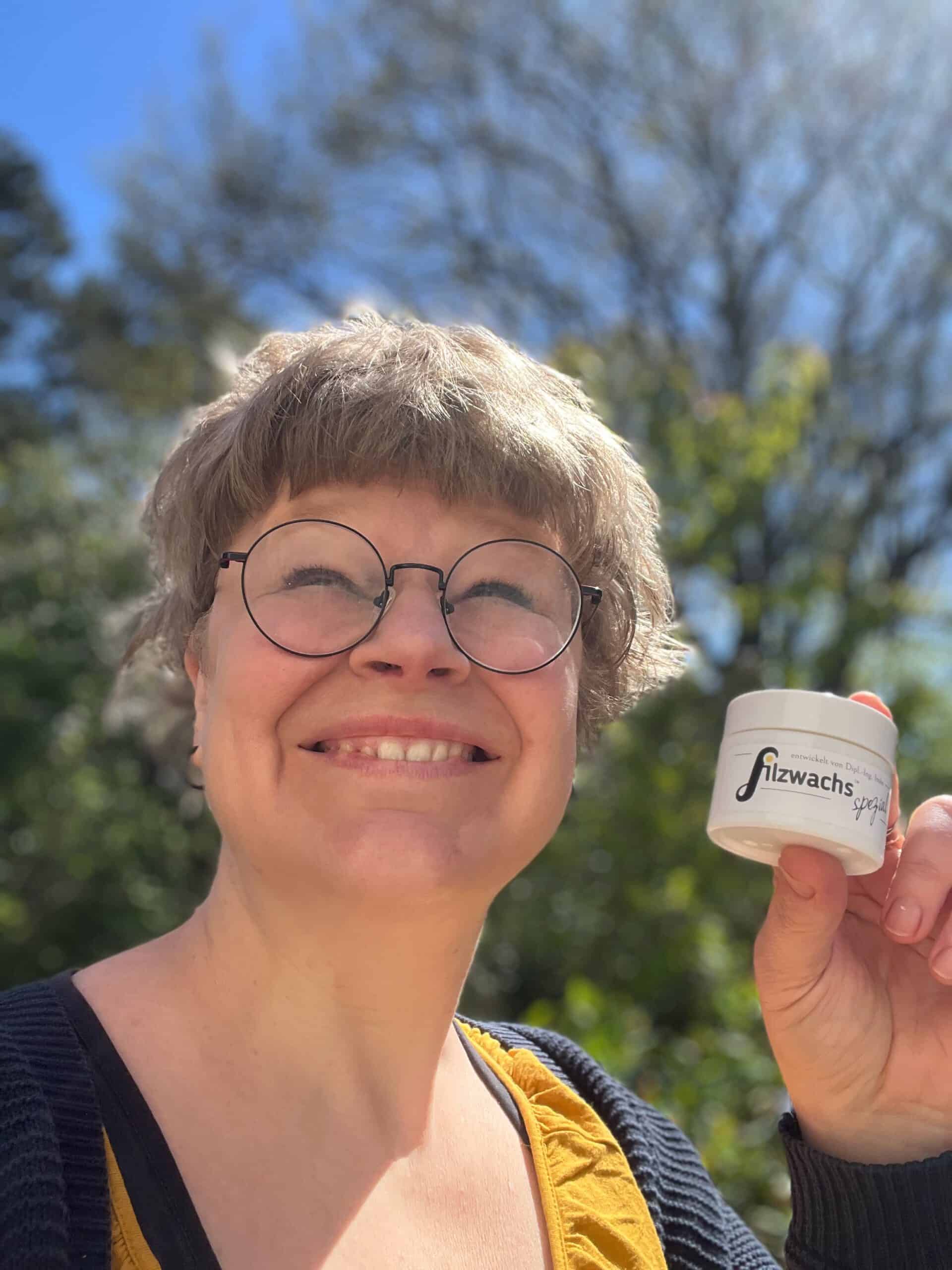 Imke, Entwicklerin von Filzwachs, hält den neuen weißen Opalglas-Tiegel Filzwachs Spezial in die Kamera – lachend im Freien vor blauem Himmel und grünen Bäumen