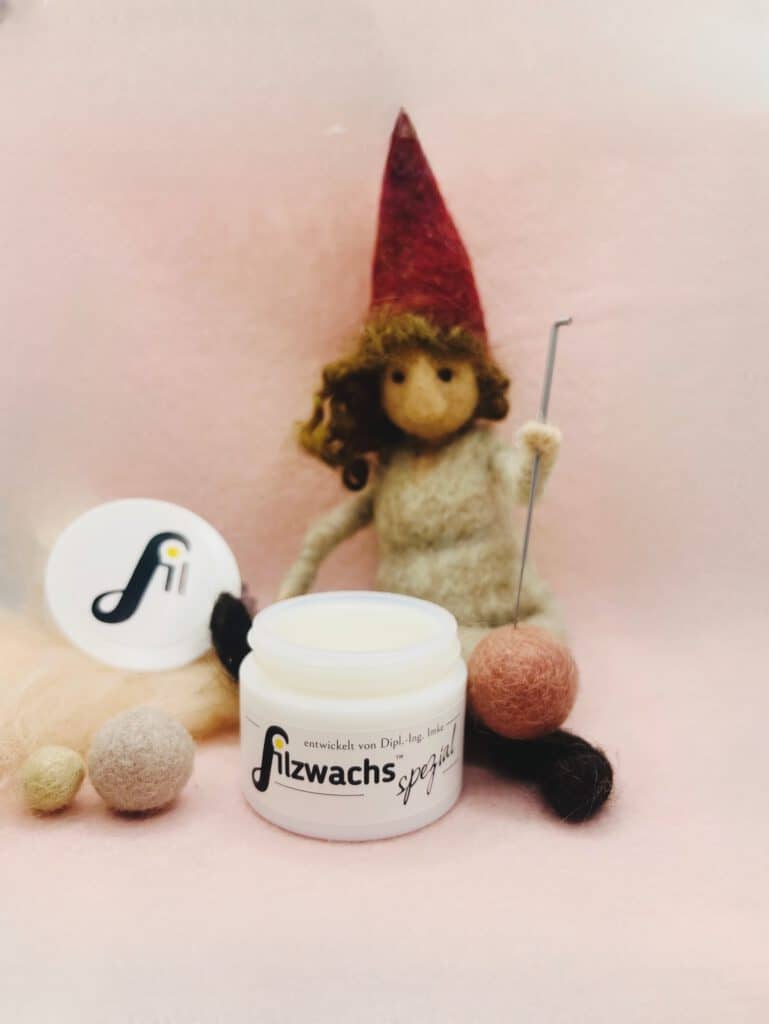 Geöffneter weißer Opalglas-Tiegel Filzwachs Spezial auf rosafarbenem Untergrund, umgeben von Wollbällchen, Rohwolle und einem handgefilzten Wichtel mit roter Mütze und Filznadel